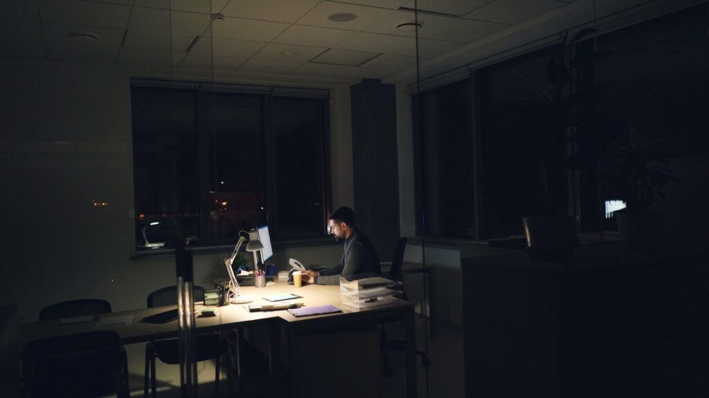 A man working alone at a desk in a dark office at night, illuminated by a desk lamp